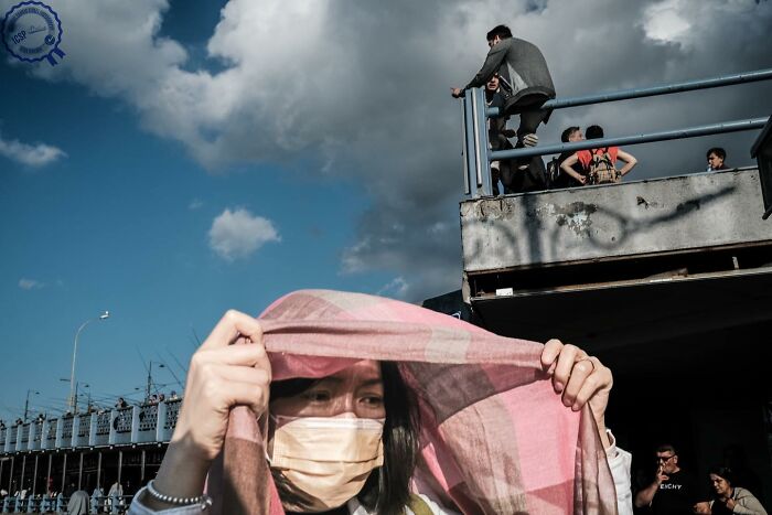 Person in a mask holds a shawl over their head, with people on a bridge in the background under a cloudy sky, street photography.