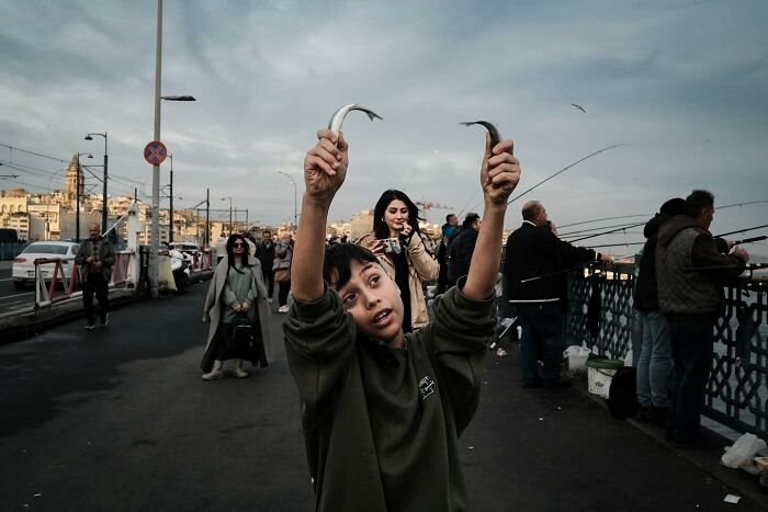A child on a bustling bridge holds two fish, showcasing Gökhan Arer's unique street photography perspective.