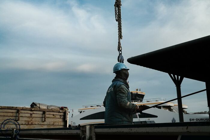 Worker in a hard hat on a dock, framed by a crane and ferry, showcasing Gökhan Arer's unique street photography.