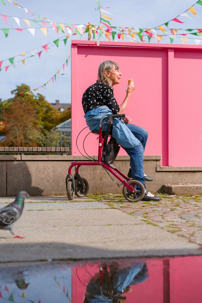 An elderly woman in Oslo enjoying ice cream on a mobility walker by a pink wall, with colorful flags overhead and a pigeon nearby.