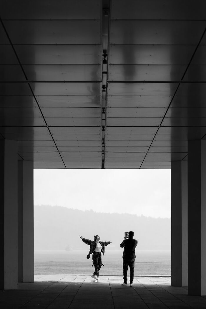 Street scene in Oslo with two people; one posing joyfully, the other photographing under a geometric canopy.