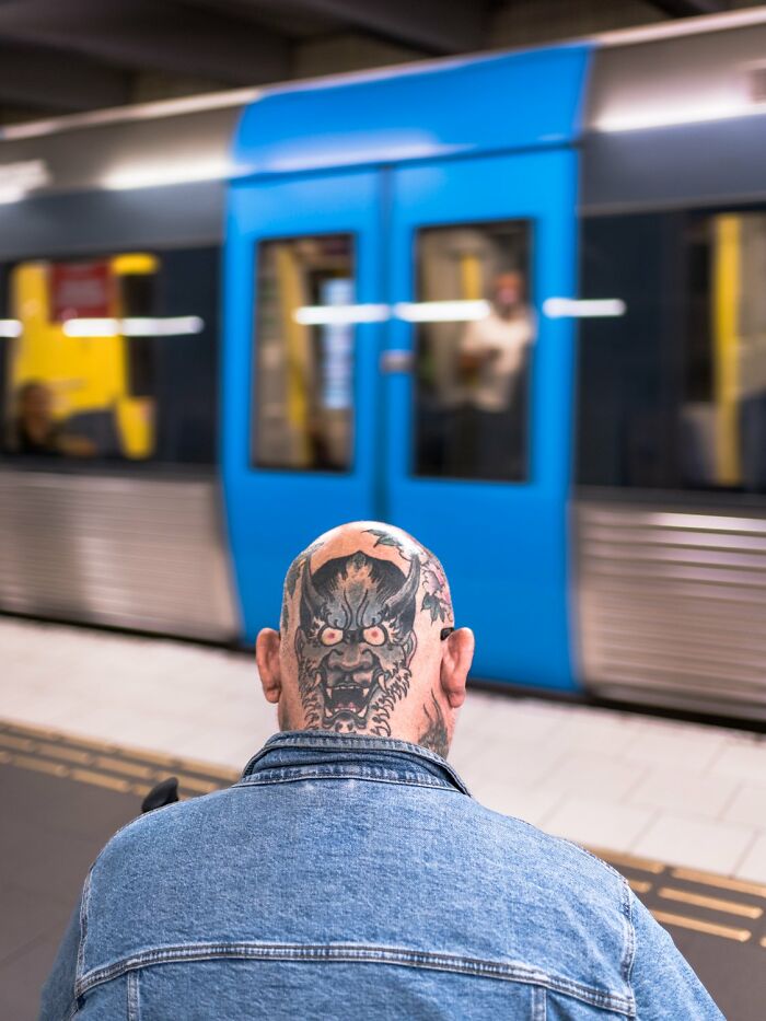 Bald man with a tattooed head in front of a blue train, capturing the essence of Oslo and Stockholm's streets.