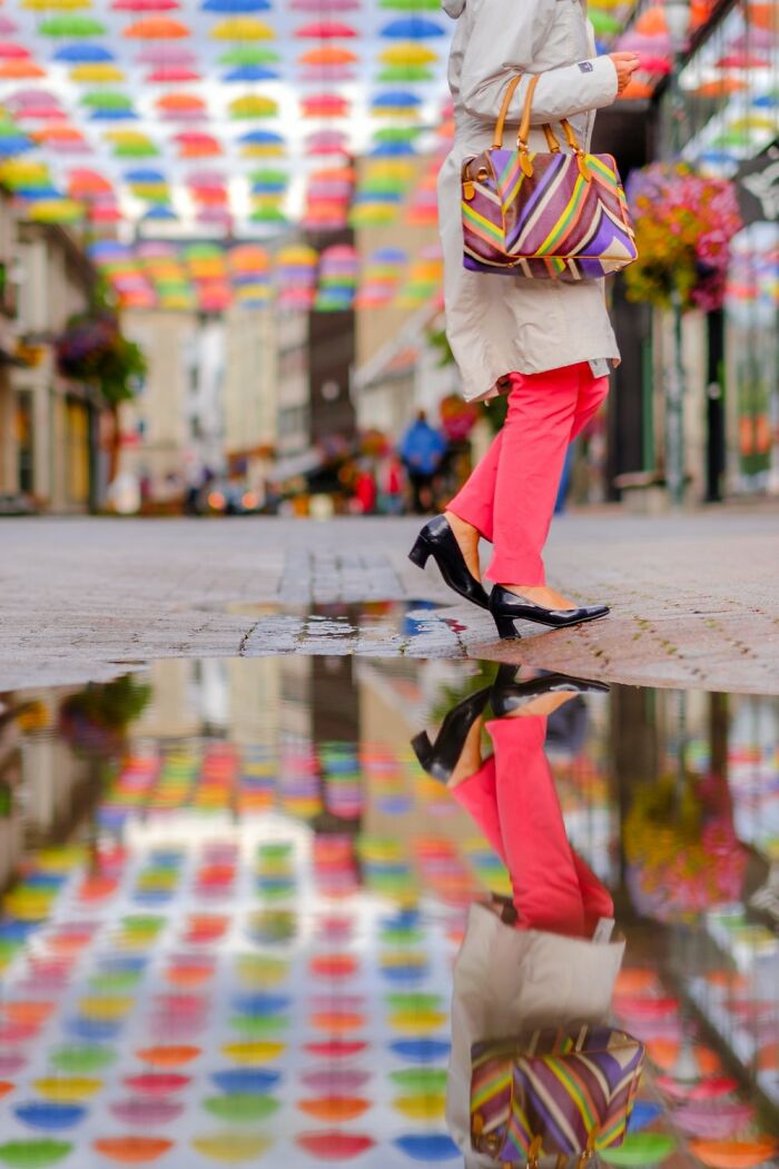 Woman walking on colorful street in Oslo, wearing pink pants and a striped bag, with umbrellas overhead reflecting in a puddle.
