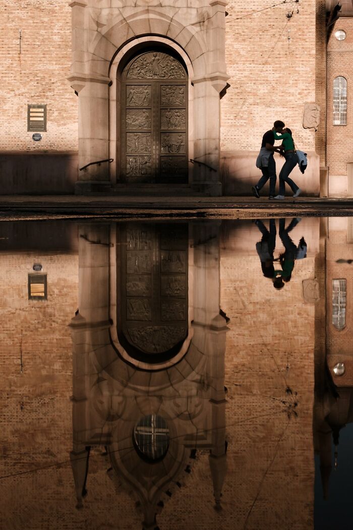 Street photo of a couple walking, reflected in water, in front of an ornate building in Oslo or Stockholm.