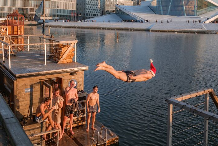 Man diving into water wearing Santa hat, captured in Oslo's urban scenery by André, with people watching from a dock.