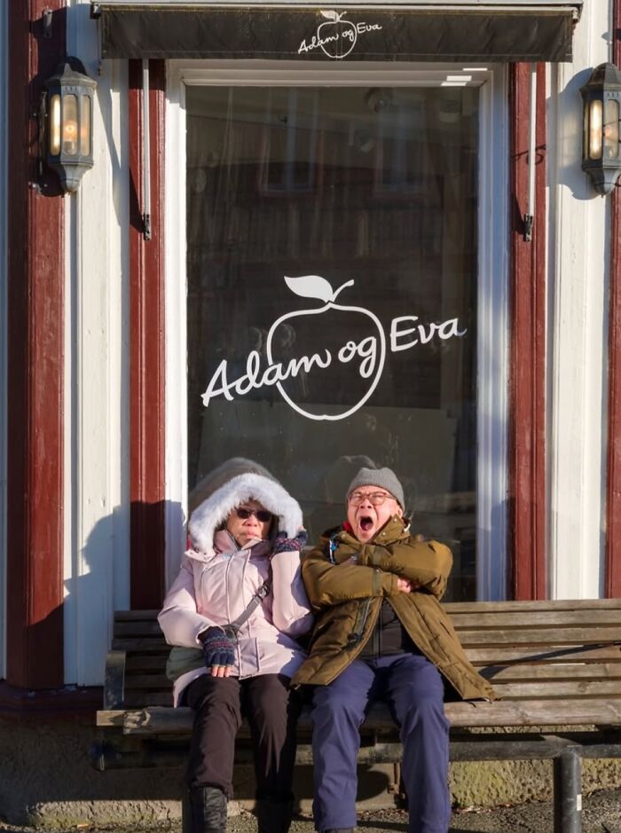 Elderly couple sitting on a bench in Oslo, in front of Adam og Eva shop window, capturing street life essence.