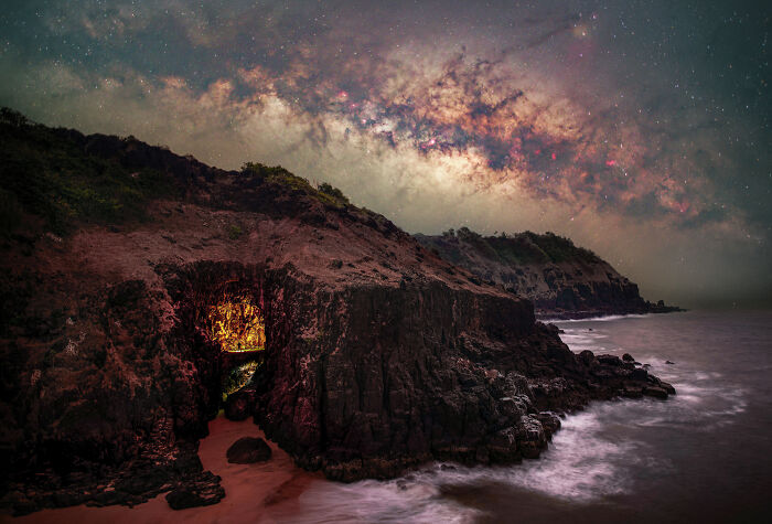 Starry night over rocky coastline in an award-winning nature photo, showcasing vibrant Milky Way and illuminated cave.