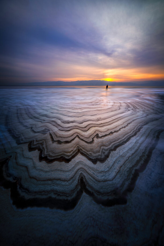 Award-winning nature photo of a person walking on layered salt flats at sunset.