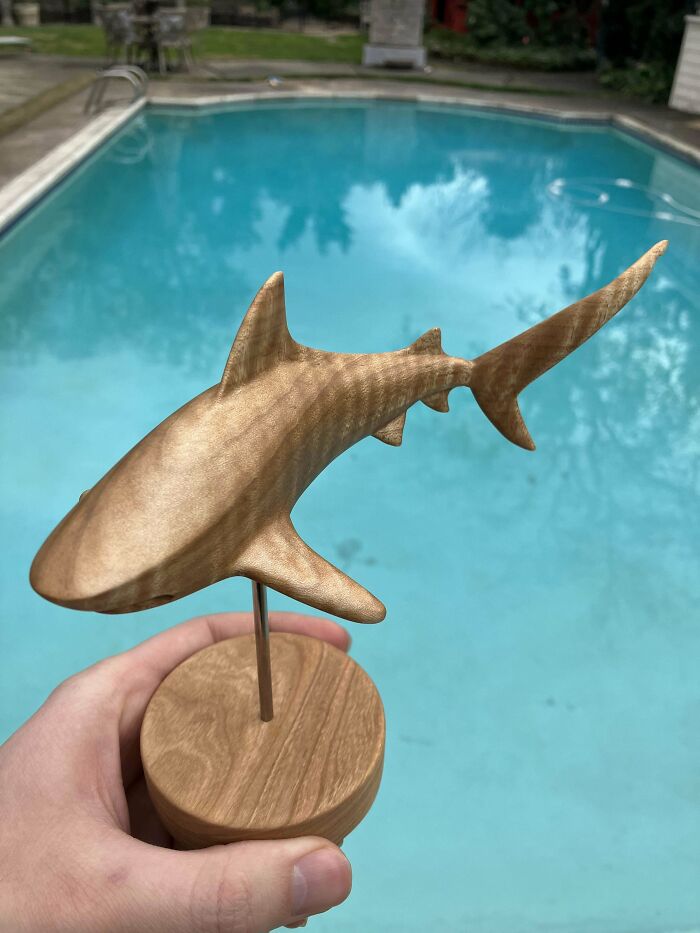 Hand holding a finely carved wooden shark sculpture with smooth finish displayed near a swimming pool, showcasing woodworking skills.