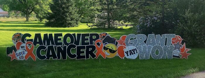 Sign fail with jumbled letters reading "Game Over Cancer" and "Grant Won" on a lawn with mixed symbols and emojis.