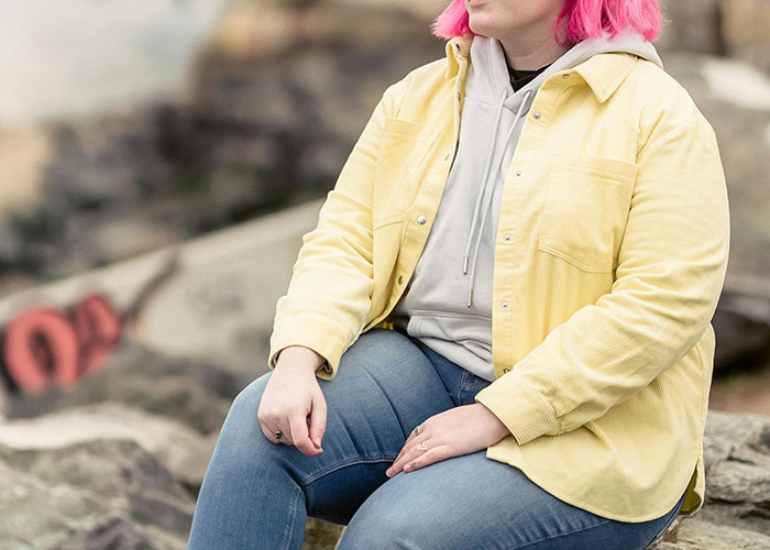 Person with pink hair in yellow jacket and jeans sitting outside, symbolizing challenges of being overweight.