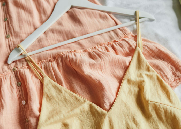 Clothes, including a yellow tank top and a peach dress on hangers, lying on a bed.