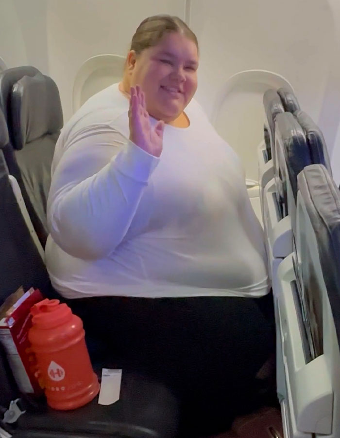 Passenger seated on a plane, waving and smiling, surrounded by travel items, amidst debate on airline discrimination fees. Passenger seated on a plane, waving and smiling, surrounded by travel items, amidst debate on airline discrimination fees.