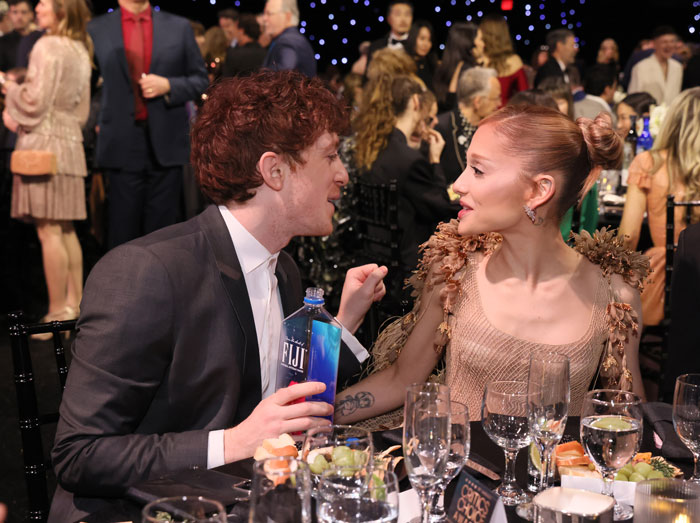 Ariana Grande at Critics Choice Awards seated at a table, engaging in conversation with a man holding a water bottle. Ariana Grande at Critics Choice Awards seated at a table, engaging in conversation with a man holding a water bottle.