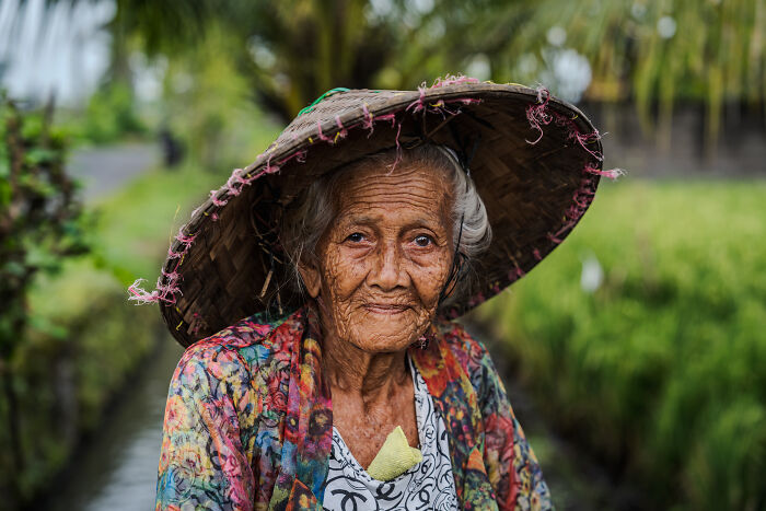 Elderly woman in a colorful outfit and straw hat, symbolizing Bali's Galungan celebration amid lush greenery.
