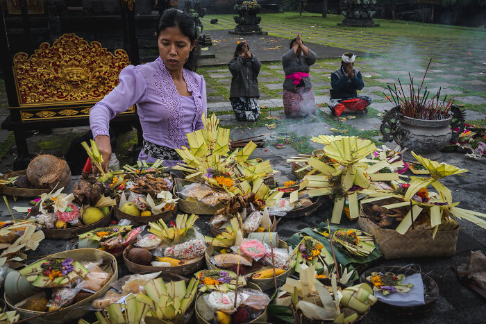 Balinese woman preparing offerings for Galungan celebration, with people praying in the background in a temple setting.