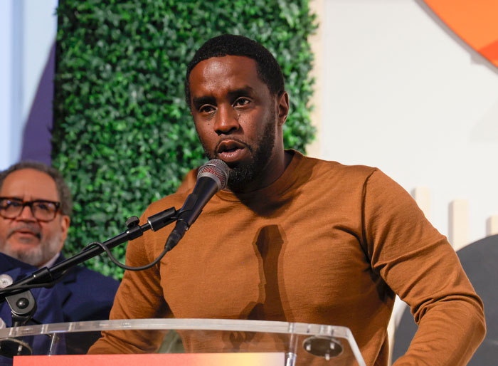 Man speaking into a microphone at an event, wearing a brown shirt, with Diddy's lawyer in the background. Man speaking into a microphone at an event, wearing a brown shirt, with Diddy's lawyer in the background.