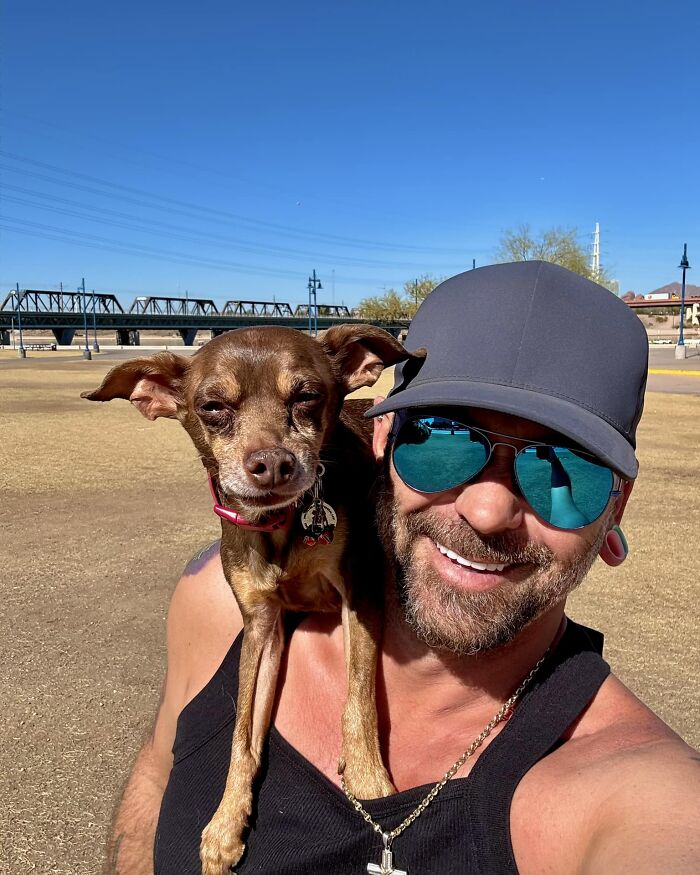 Man smiling with lost dog on his shoulder, wearing sunglasses and cap, outdoors with blue sky background. Man smiling with lost dog on his shoulder, wearing sunglasses and cap, outdoors with blue sky background.