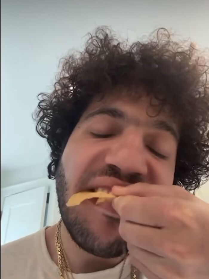 A person with curly hair biting into a chip, wearing a white shirt and gold chains, related to Valentine's Day gift topic. A person with curly hair biting into a chip, wearing a white shirt and gold chains, related to Valentine's Day gift topic.