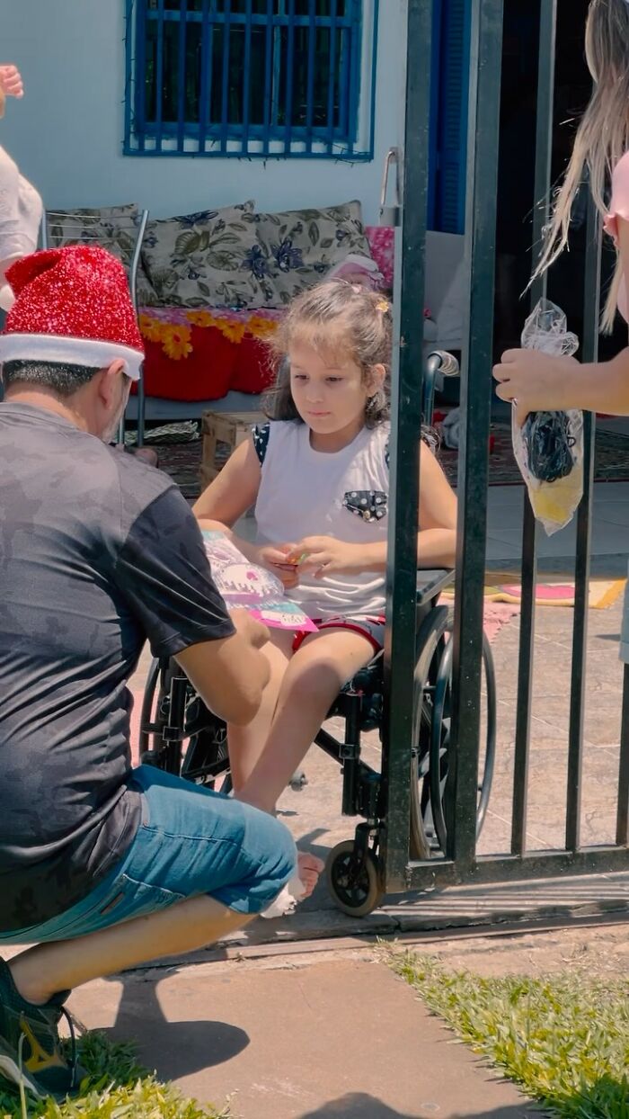 Man in Santa hat kneeling by a young girl in a wheelchair near a gate, handing her a book. Man in Santa hat kneeling by a young girl in a wheelchair near a gate, handing her a book.