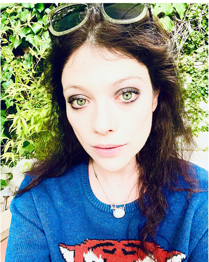 A woman with green eyes and dark hair, wearing a blue sweater and sunglasses, poses with greenery in the background. A woman with green eyes and dark hair, wearing a blue sweater and sunglasses, poses with greenery in the background.