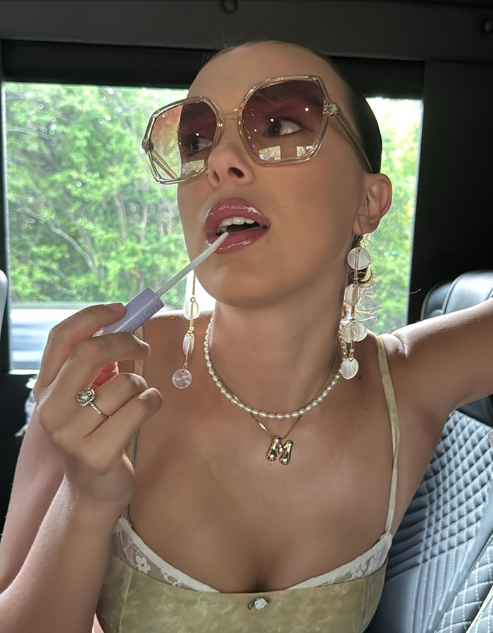 Millie Bobby Brown applies lip gloss in a car, wearing oversized sunglasses and jewelry. Millie Bobby Brown applies lip gloss in a car, wearing oversized sunglasses and jewelry.