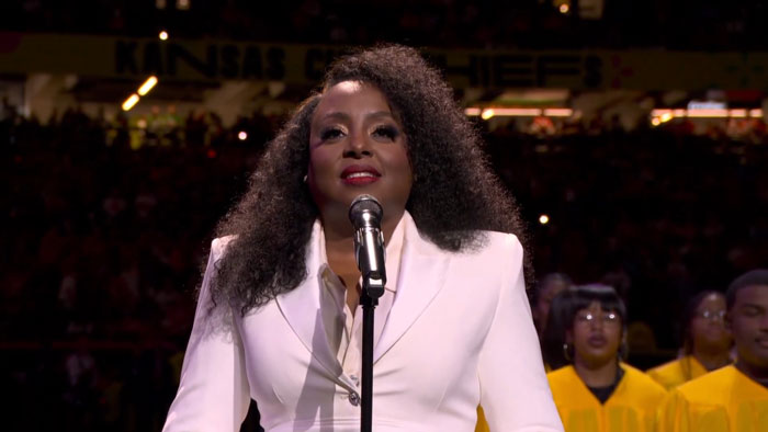 Singer performing national anthem at Super Bowl LIX, wearing a white suit, with audience in the background. Singer performing national anthem at Super Bowl LIX, wearing a white suit, with audience in the background.