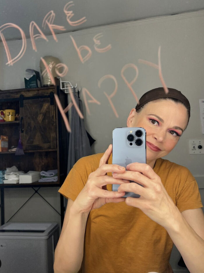 Person in a dressing room mirror, smiling and taking a selfie. "Dare to be happy" written on the mirror. Person in a dressing room mirror, smiling and taking a selfie. "Dare to be happy" written on the mirror.