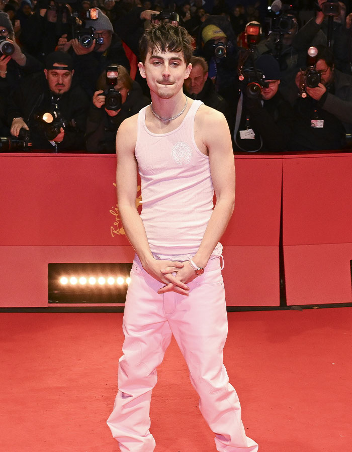 Man in white tank top and pants at Valentine’s Day red carpet, surrounded by photographers. Man in white tank top and pants at Valentine’s Day red carpet, surrounded by photographers.
