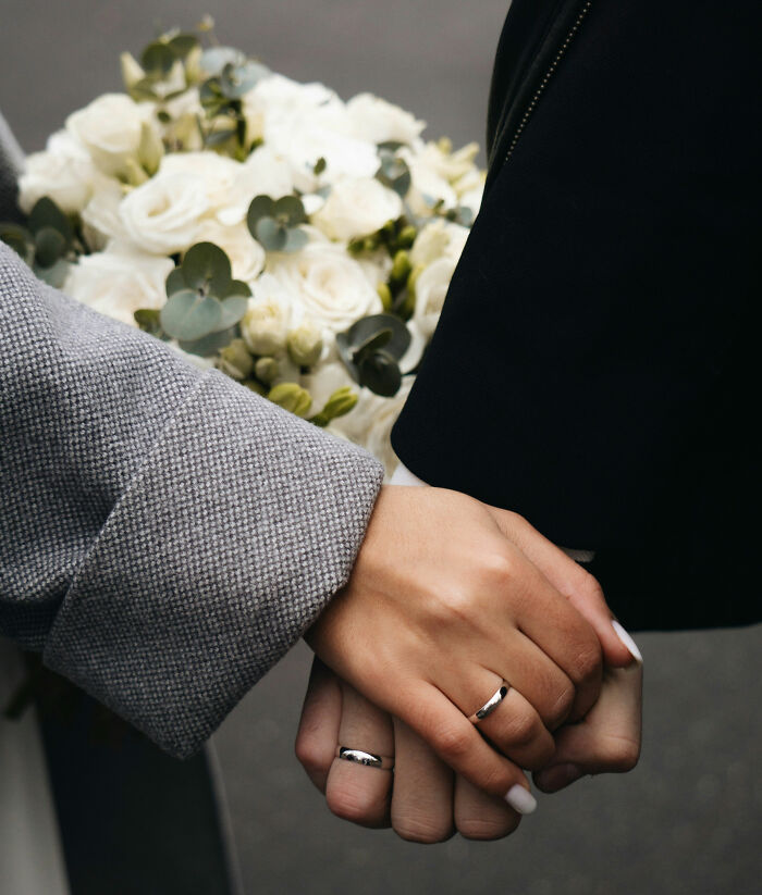 Hands of women wearing rings, holding hands affectionately with a bouquet of white roses in the background.
