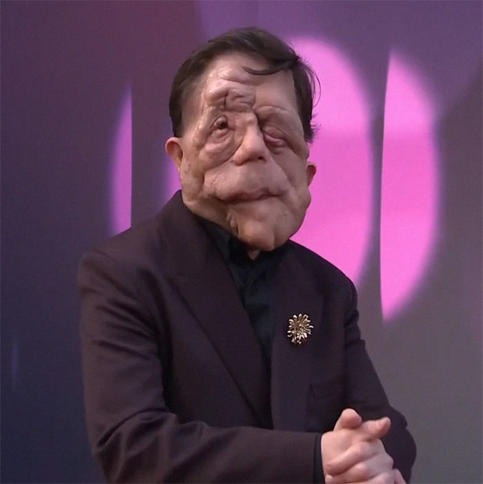 Person in a dark suit and brooch stands against a purple background, embodying resilience despite "anti-trans" claims. Person in a dark suit and brooch stands against a purple background, embodying resilience despite "anti-trans" claims.