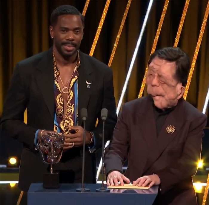 Two men standing at a podium, man on right holding a paper. Event lighting in the background. Two men standing at a podium, man on right holding a paper. Event lighting in the background.
