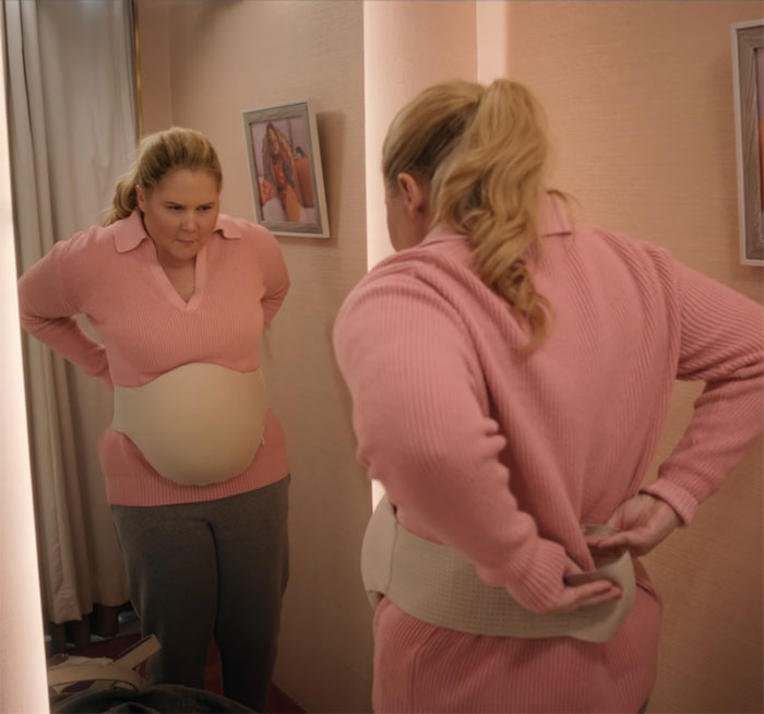 Amy Schumer in a pink sweater looks in the mirror, adjusting a waist cincher, related to her new comedy. Amy Schumer in a pink sweater looks in the mirror, adjusting a waist cincher, related to her new comedy.