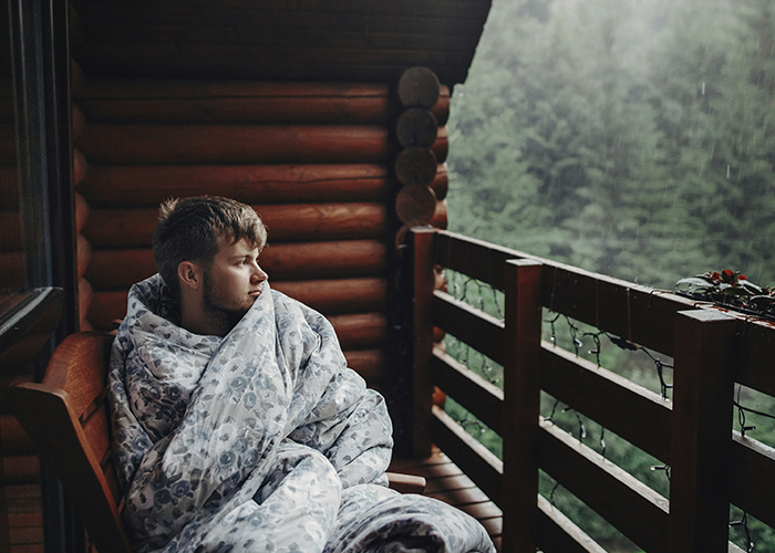 A man wrapped in a blanket sits on a wooden cabin porch, gazing at a rainy forest view. A man wrapped in a blanket sits on a wooden cabin porch, gazing at a rainy forest view.