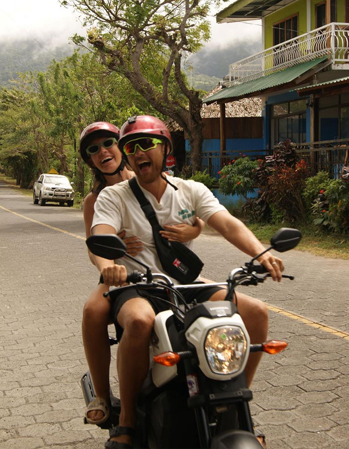 Couple riding a motorbike in Thailand, wearing helmets and smiling on an empty road with scenic greenery in the background. Couple riding a motorbike in Thailand, wearing helmets and smiling on an empty road with scenic greenery in the background.