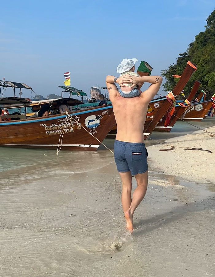 A person carrying a baby at a beach in Thailand, near wooden boats, under a clear sky. A person carrying a baby at a beach in Thailand, near wooden boats, under a clear sky.