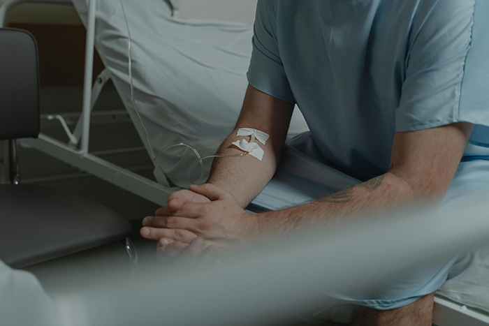 Man in hospital attire sitting beside a bed, with an IV bandage on his arm. Man in hospital attire sitting beside a bed, with an IV bandage on his arm.