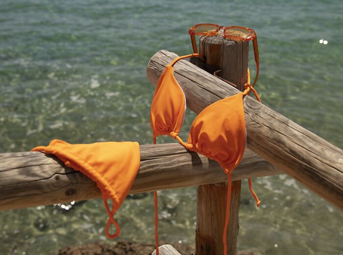 Orange bikini and sunglasses on a wooden fence by the ocean, highlighting recent fashion trends people hate.
