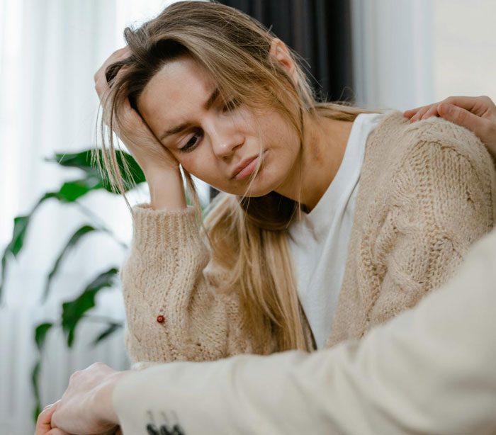 Woman looking distressed, wearing a beige sweater, sitting indoors with a hand on her shoulder, related to a friend's marriage lie. Woman looking distressed, wearing a beige sweater, sitting indoors with a hand on her shoulder, related to a friend's marriage lie.