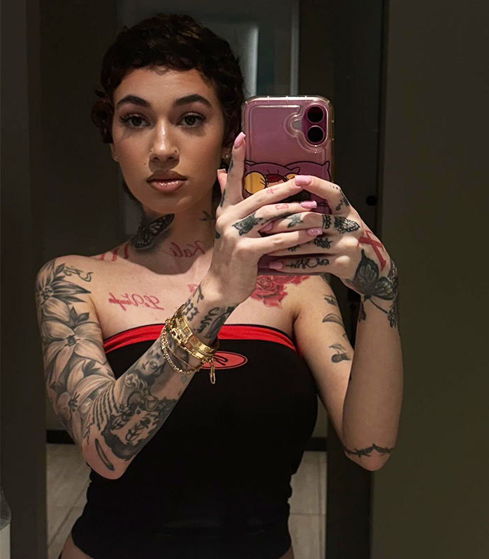 Young woman with tattoos taking a selfie, holding a pink phone, wearing a black top and gold bracelets. Young woman with tattoos taking a selfie, holding a pink phone, wearing a black top and gold bracelets.