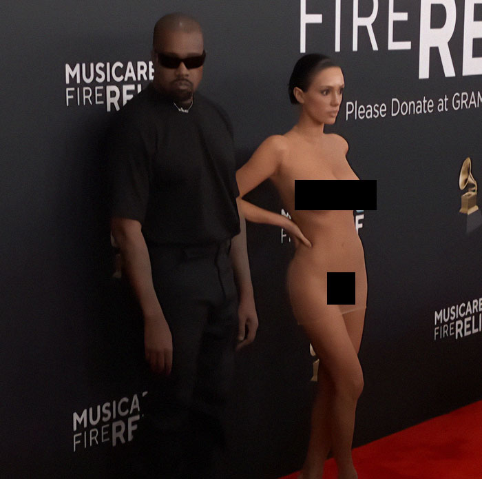 A person in sunglasses stands next to Bianca Censori in a controversial outfit at a Grammys event. A person in sunglasses stands next to Bianca Censori in a controversial outfit at a Grammys event.