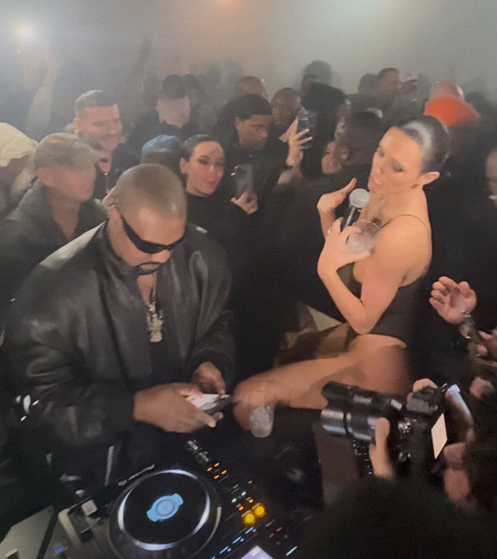 Bianca Censori at afterparty, holding a microphone, surrounded by a crowd in a dimly lit room. Bianca Censori at afterparty, holding a microphone, surrounded by a crowd in a dimly lit room.