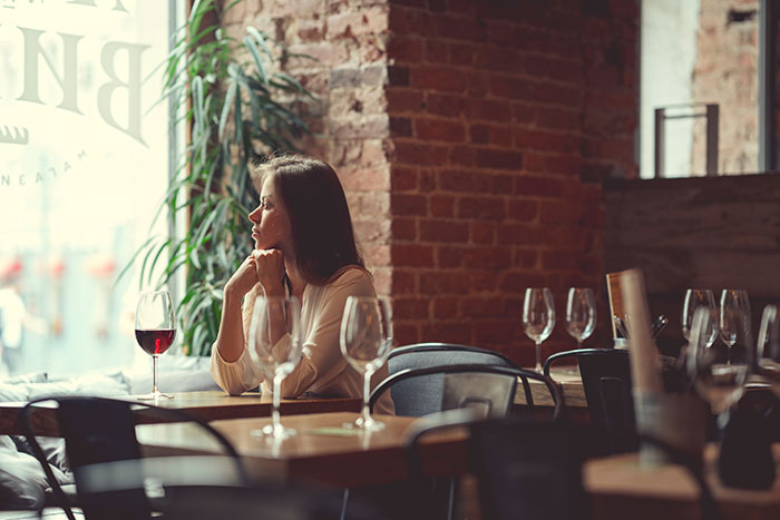 Woman sitting alone at a restaurant table with empty chairs, looking thoughtful, with a glass of red wine in front of her. Woman sitting alone at a restaurant table with empty chairs, looking thoughtful, with a glass of red wine in front of her.