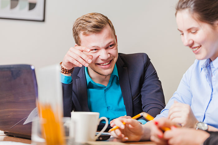 Man in a blue shirt laughing in an office meeting, sitting next to a coworker with a laptop and coffee mug. Man in a blue shirt laughing in an office meeting, sitting next to a coworker with a laptop and coffee mug.