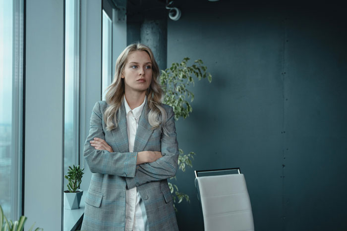 A woman in a business suit standing by an office window, arms crossed, representing a heroic coworker scenario. A woman in a business suit standing by an office window, arms crossed, representing a heroic coworker scenario.