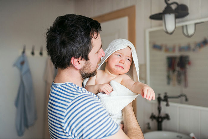Father struggles to calm crying infant in towel, reflecting drama over parental responsibilities. Father struggles to calm crying infant in towel, reflecting drama over parental responsibilities.