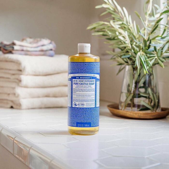 Dr. Bronner's multi-functional Castile soap bottle on a tiled countertop with folded towels and a potted plant nearby.