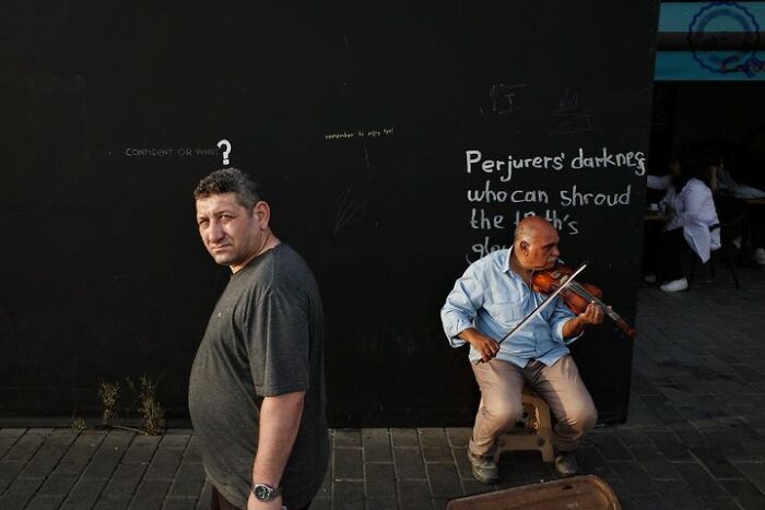Man walking past a violinist on a street, graffiti visible, showcasing Gökhan Arer's unique street photography perspective.