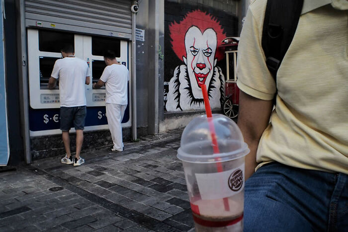 Street artist's view: two men at ATMs beside a Pennywise mural, with a blurred drink in the foreground.