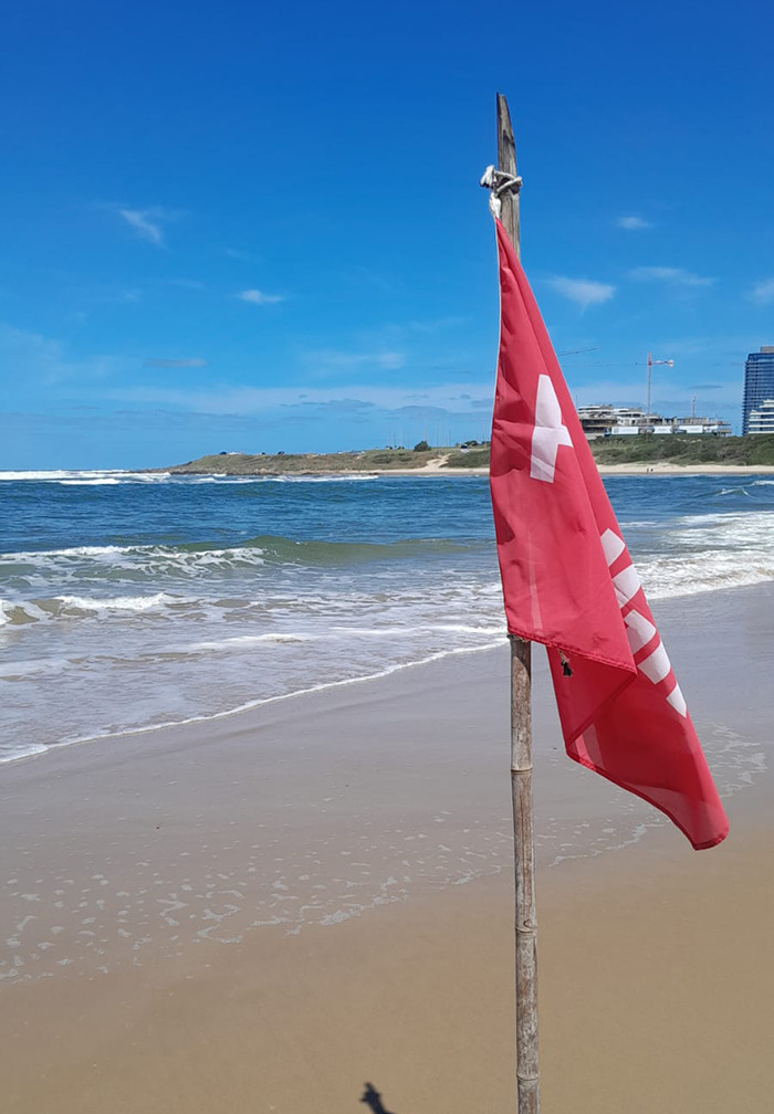 Beach scene with a red lifeguard flag on a pole near the shoreline under a clear blue sky. Beach scene with a red lifeguard flag on a pole near the shoreline under a clear blue sky.
